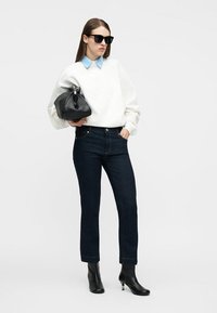 Γυναίκα με μαύρα γυαλιά ηλίου, λευκό πουλόβερ με μπλε γιακά, σκούρο τζιν, μαύρα μποτάκια μέχρι τον αστράγαλο, κρατώντας μαύρη τσάντα σε ένα απλό φόντο.