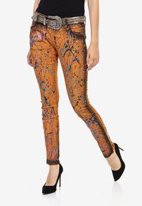 Jean skinny en denim orange avec un motif éclaboussé en bleu et rose. Présente des clous métalliques le long des poches et une ceinture décorative ornée de strass.