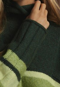 Strickpullover mit dunkelgrünen und hellgrünen Streifenmustern, einem hohen Kragen und gerippten Bündchen, hergestellt aus weichem, strukturiertem Stoff.