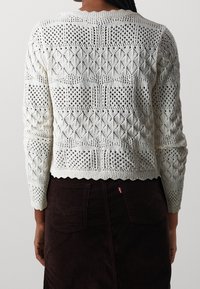 Vit stickad tröja med intrikata mönster, med rund hals och ribbade kanter. Designen inkluderar diamant- och hålmönster.