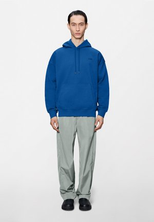 Man standing wearing a blue hoodie, light gray loose pants, and black slip-on shoes against a plain white background.