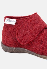 Rode wollen slipper met een gestructureerde zool en een klittenbandsluiting, met een wit label met "Gottstein" aan de zijkant.