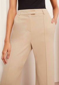 Beige weit geschnittene Hose aus strukturiertem Stoff, mit Fronttaschen und taillierter Taille mit Gürtelschlaufen. Sauberes, modernes Design.