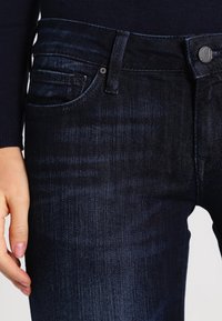 Jeans de mezclilla azul oscuro con un corte ajustado, diseño de cinco bolsillos y una textura sutil. Cierre de botón metálico con un acento de marca.