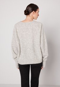 Tumejuustega naine, kes kannab lahtist helehallist kootud kampsunit ja liibuvaid musti pükse, seisab tavalise heleda tausta ees.