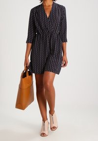 Robe portefeuille bleu marine à manches trois quarts et taille cintrée, ornée d'un motif abstrait blanc. Portée avec des bottines ouvertes beiges et un sac fourre-tout marron.