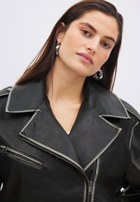 Veste en simili cuir noir avec revers larges, détails en fermeture éclair argentée et parement contrasté. Portée avec des grandes boucles d'oreilles en argent.