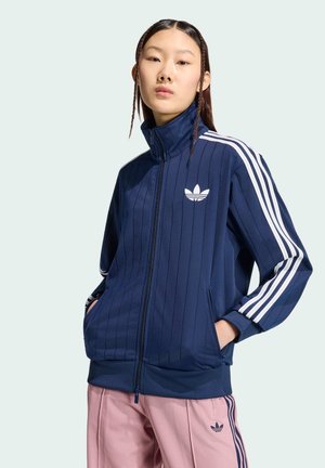 Young person with long hair in braids wears a navy blue Adidas zip-up jacket and pink Adidas track pants, standing with hands in pockets.