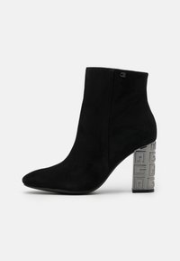 Botín de ante negro hasta el tobillo con punta afilada y un tacón de bloque metálico que presenta un patrón geométrico. Textura suave y diseño elegante.