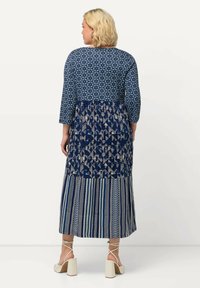 Abito lungo con maniche a tre quarti, con un motivo geometrico blu e crema, design a strati e orlo svasato. Indossato con sandali con tacco a blocco.