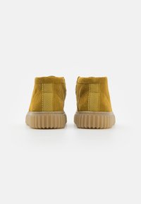 Gelbe Wildleder-Ankle Boots mit gerippten Gummisohlen, von hinten auf einem weißen Hintergrund gezeigt.