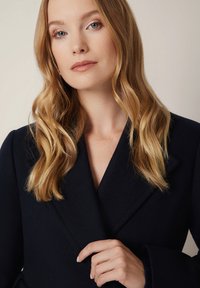 Granatowy wełniany płaszcz z klapami, o miękkiej teksturze i długich rękawach. Osoba na zdjęciu ma falujące włosy i delikatnie trzyma płaszcz.