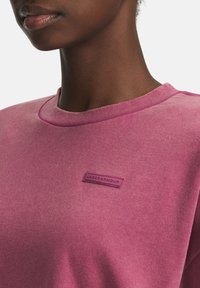 Close-up van een persoon die een vervaagde roze sweatshirt met ronde hals draagt, met een klein Under Armour-logo op de borst.