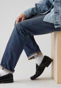 Jeans de mezclilla azul oscuro con los puños doblados, combinados con mocasines negros y calcetines blancos. La mezclilla muestra una sutil textura y detalle de costura.