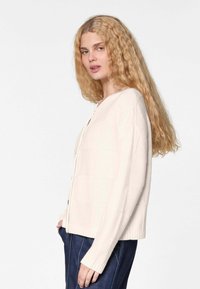 Cremefarbener Cardigan aus weichem Strickstoff, mit V-Ausschnitt, Knopfverschluss und gerippten Ärmeln; locker geschnitten mit einer kastigen Form.