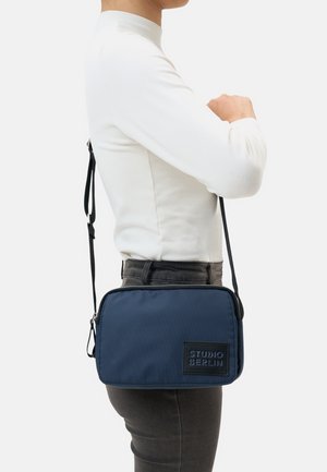 Navy blue crossbody bag with textured fabric, zip closure, and a prominent black logo patch. Adjustable strap and rectangular shape.