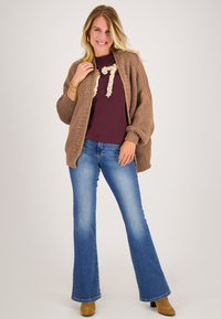 Gebreide brown cardigan, bordeauxhemd met textuurdetails, blauwe uitlopende jeans en tan enkellaarsjes; losse pasvorm en casual ontwerp.