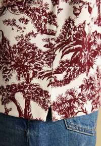Bavlnená košeľa s vínovým kvetinovým vzorom na bielom pozadí, s mierne zakriveným lemom a jemnou textúrou, kombinovaná s modrými denimovými džínami.