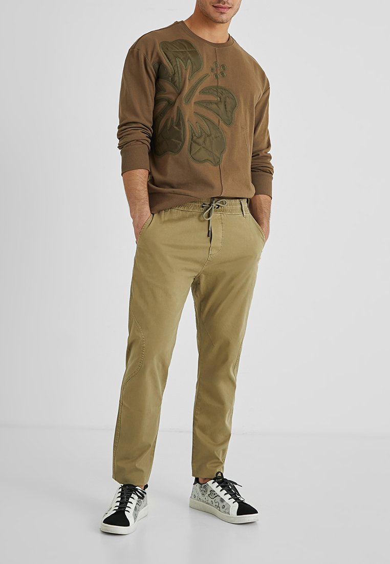 Brauner Sweatshirt mit Blättermuster, kombiniert mit beigen Hosen mit elastischem Bund und Sneakers mit schwarzen Akzenten.