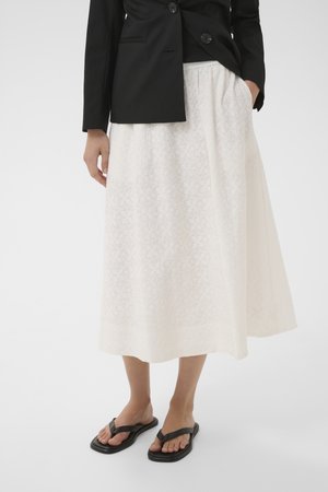 Kvinde iført hvid tekstureret midi-nederdel, sort blazer og sorte thongsandaler, stående med den ene hånd i lommen på nederdelen.