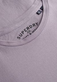 T-shirt en coton lavande avec un col rond. Présente une texture subtile, des coutures visibles et une étiquette de marque imprimée à l'intérieur du col.