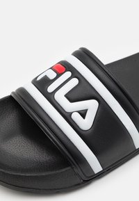 Svart slidesandal med en tjock rem som har ett vitt streck och en upphöjd vit logga. Texturerad svart gummisula med lätt krökning.