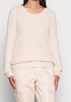 Femme portant un pull en maille texturé crème clair à manches longues et un pantalon cargo beige avec poches avant.