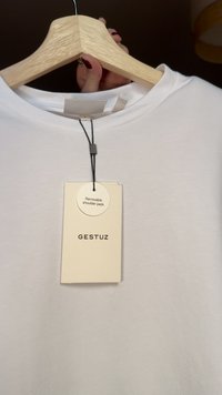 Maglietta bianca esposta su una gruccia di legno, con un'etichetta che indica "spalline removibili" con testo nero e forma rotonda.