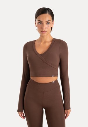 Kvinde med kort mørkt hår iført matchende brun langærmet crop top og højtaljede leggings, stående foran en ensfarvet lys baggrund.