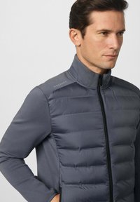 Hombre vestido con una chaqueta gris acolchada con cremallera, con el frente acolchado y mangas lisas, mirando hacia la derecha frente a un fondo sencillo.