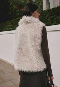 Weißweste aus Kunstpelz mit hohem Kragen, flauschiger Textur und ärmellosem Design, getragen über einem dunkelbraunen Langarmshirt.