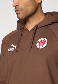 Sudadera marrón con cordones, con un parche de logo en rojo y blanco, y el texto "KING" en blanco en el lado izquierdo del pecho. Diseño atlético.