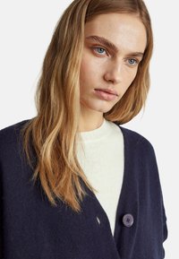 Tmavě námořnický cardigan s designem do V, který má jeden fialový knoflík a měkkou texturu, vrstvený přes svetřík světlé barvy.