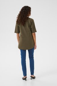 Olive green oversize koszula z krótkimi rękawami, połączona z dopasowanymi niebieskimi dżinsami i czarnymi sandałami na obcasie, widok z tyłu.