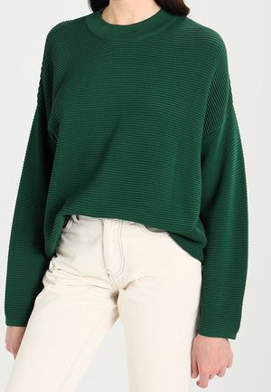 Femme portant un pull côtelé vert foncé à manches longues rentré dans un pantalon taille haute écru, debout devant un fond uni.