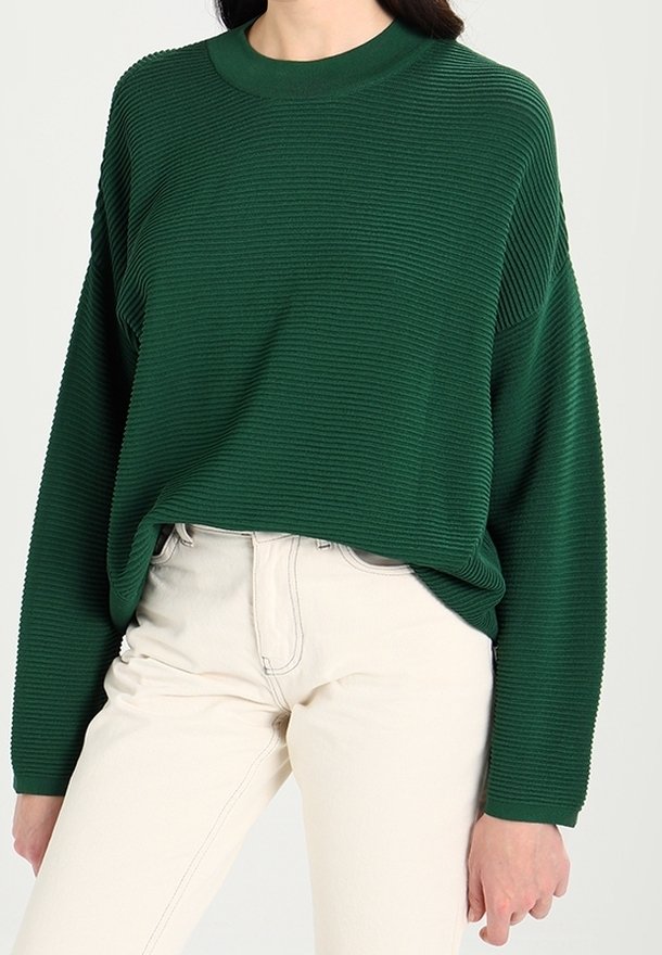 Femme portant un pull côtelé vert foncé à manches longues rentré dans un pantalon taille haute écru, debout devant un fond uni.