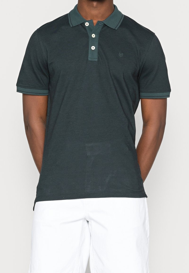 Polo vert foncé à manches courtes avec deux boutons blancs porté par une personne, assorti à un short blanc, fond uni.