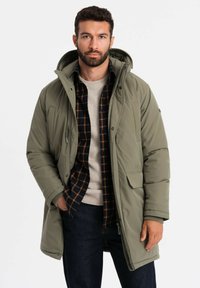 Olivgrüner Parka mit Kapuze, mit Reißverschluss und Knopfverschluss, zwei Vordertaschen, getragen über einem karierten Hemd und einem beigen Pullover.