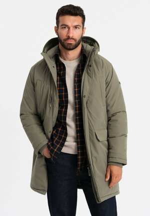 Parka verde oliva con capucha, con cierre de cremallera y botones, dos bolsillos frontales, llevada sobre una camisa de cuadros y un suéter beige.