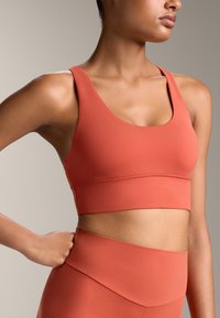 Rust narancsszínű sportmelltartó, sima, rugalmas anyagból készült. Széles, lekerekített nyakkivágással és diszkrét varrási részletekkel rendelkezik.