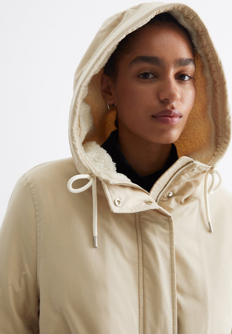 Beige Kapuzenjacke mit einem weichen, strukturierten Futter, verstellbaren Zugbändern und einem Reißverschluss vorne mit Metallbeschlägen.