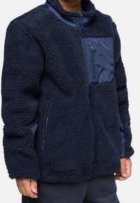 Enfant portant une veste en sherpa polaire bleu marine avec des poches zippées sur la poitrine et les côtés, debout devant un fond uni.