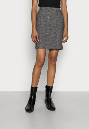 Femme portant un haut noir, une minijupe à carreaux gris et des bottines noires, debout devant un fond blanc uni.