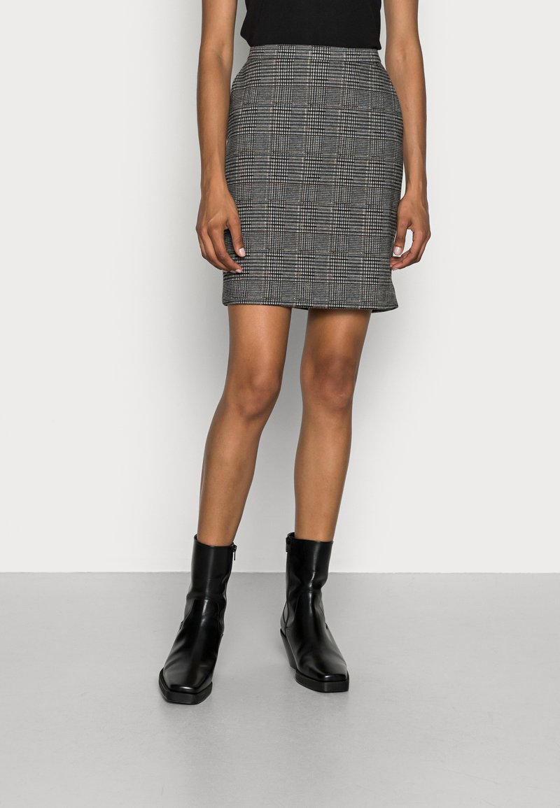 Femme portant un haut noir, une minijupe à carreaux gris et des bottines noires, debout devant un fond blanc uni.