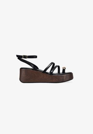 Sandalia negra de plataforma con suela de cuña de madera, tira con hebilla en el tobillo, múltiples tiras finas y anillo para el dedo del pie con una cuenta dorada decorativa.
