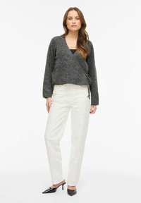 Pilkas megztinis su ilgomis rankovėmis ir šonine surišimo detale, derinamas su baltomis tiesiomis kelnėmis ir juodomis aukštakulnėmis mule batais.