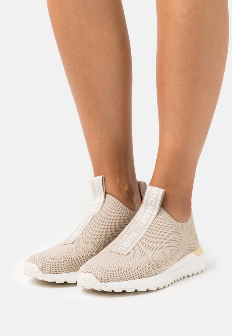 Sneaker beige senza lace realizzate in materiale a maglia traspirante con una finitura testurizzata, dotate di una cintura con logo bianco e accenti della suola in contrasto.