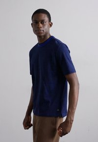 Marineblå kortermet T-skjorte med rund hals, strukturert stoff og en liten logo detalj nær kanten, iført med lysebrune bukser.