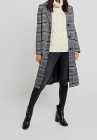 Rutig lang frakke i blå, grå og hvide nuancer. Cremefarvet kabelstrik sweater, sorte skinny jeans og sorte ankelstøvler med hæl.