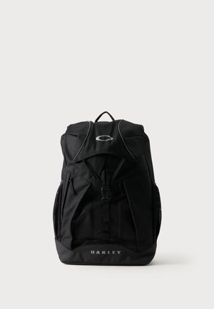 Mochila negra Oakley con hebilla frontal, múltiples compartimentos con cremallera y asa superior sobre fondo liso claro.